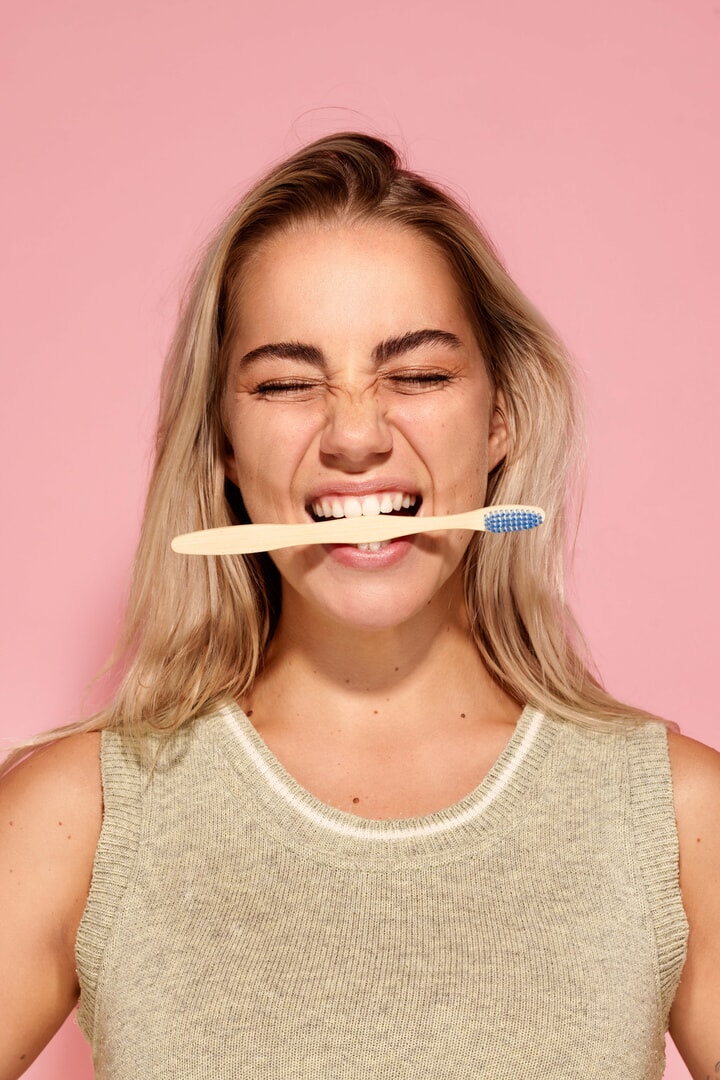 Vrouw glimlacht terwijl ze speels op een houten tandenborstel bijt, met een frisse uitstraling tegen een roze achtergrond – symbool voor een gezonde ochtendroutine.