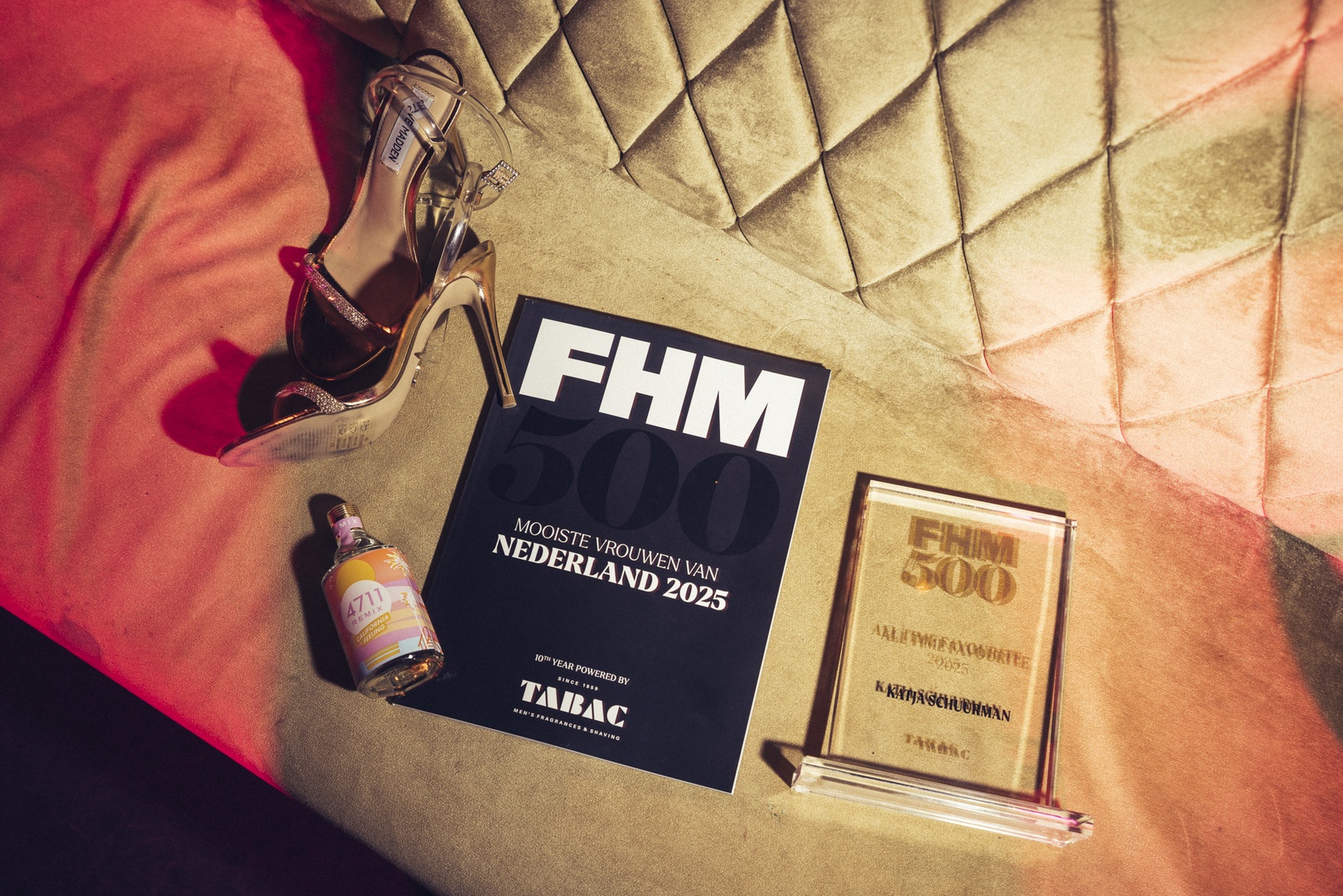 Een glamoureuze setting met gouden high heels, een flesje geurende 4711 cologne, een FHM 500 magazine met de titel “Mooiste vrouwen van Nederland 2025”, en een glazen award met de inscriptie “FHM 500 All Time Favorite 2025 – KATJA SCHUURMANS” op een fluwelen bank met gouden stiksels.