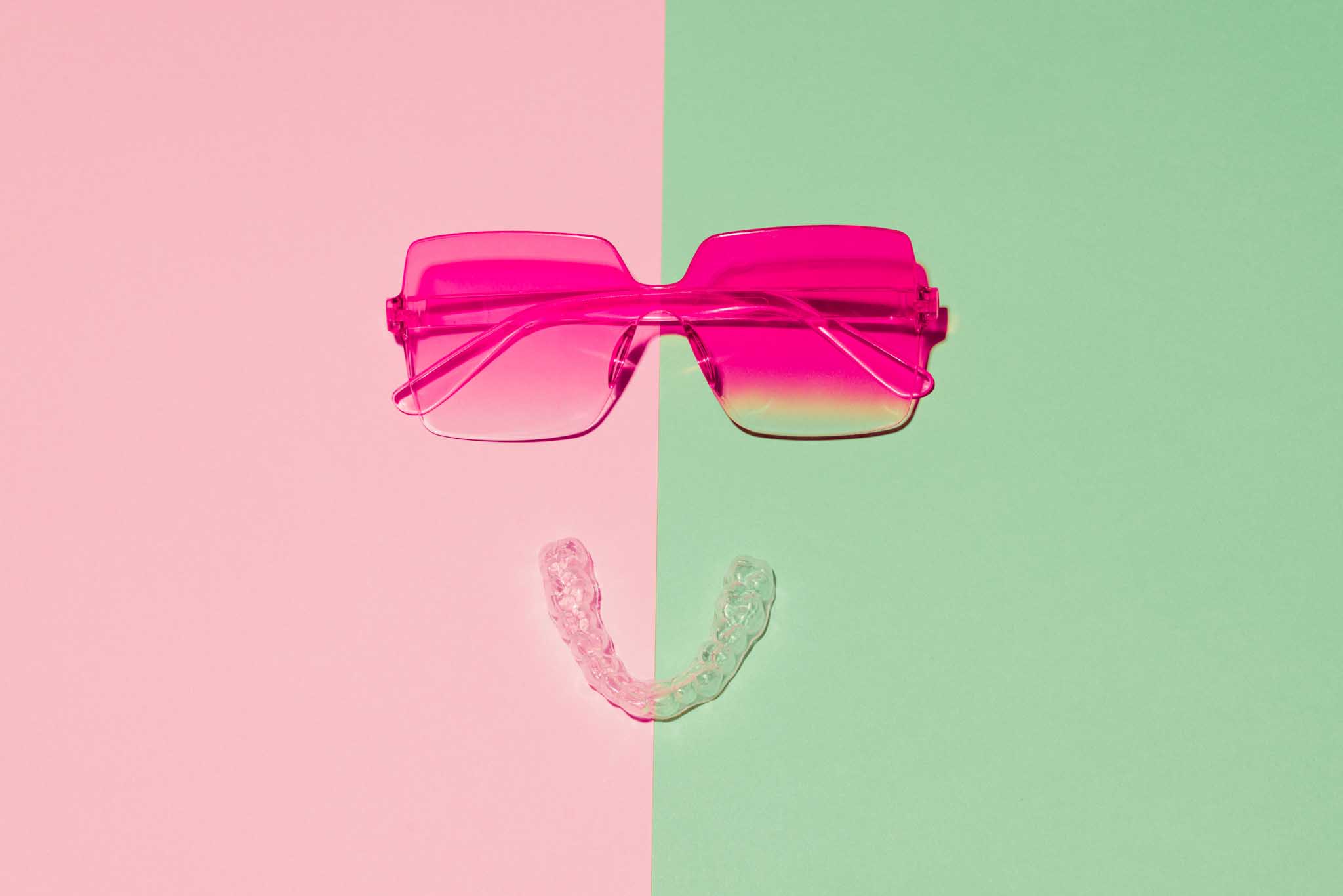 Vrolijke en speelse compositie van een felroze zonnebril en een doorzichtige aligner op een split background in pastelroze en mintgroen, waardoor het geheel lijkt op een lachend gezicht.